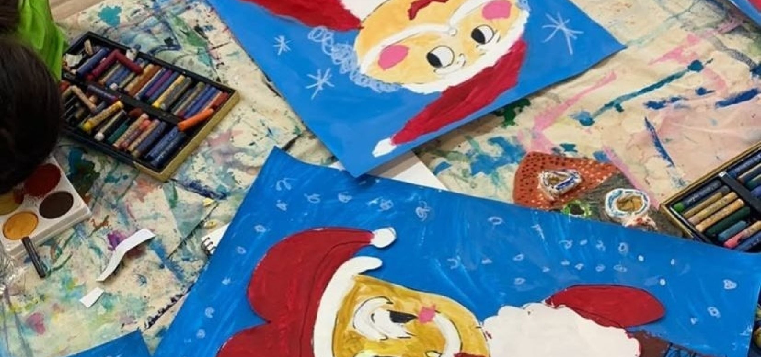 Children painting colorful Christmas-themed pictures of Santa Claus on blue paper at a craft table with art supplies