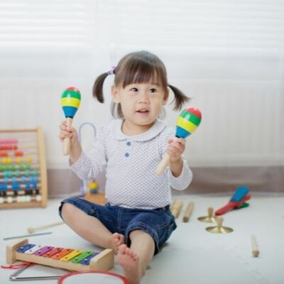 young girl of approximately two years old wearing a white top and jeans sitting on a white carpet floor holding a colourful maracas in each hand and surrounded by various other colourful instruments including a guitar and xylophone.