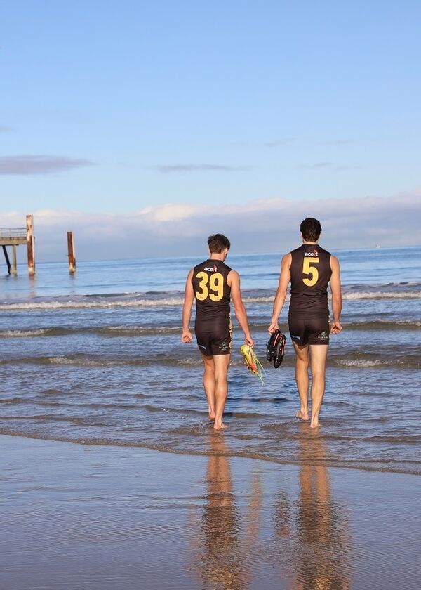 Two people walk into the ocean with the Glenelg Jetty in the background.