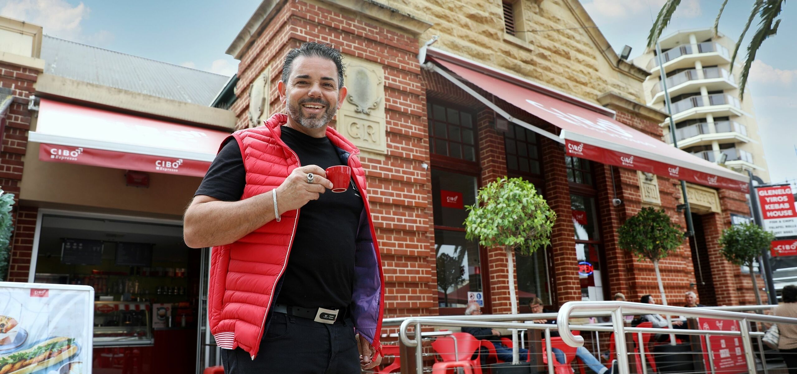 Man holding red espresso cup wearing a red puffer vest, in front of the cibo glenelg building