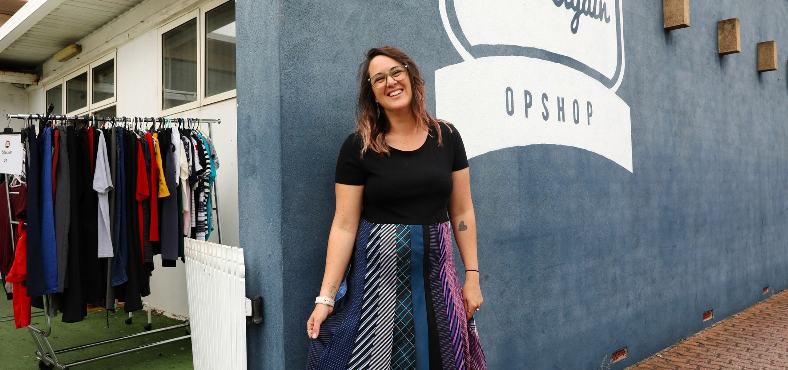 Woman standing in a skirt made of ties in front of a grey wall that says "worn again op shop"