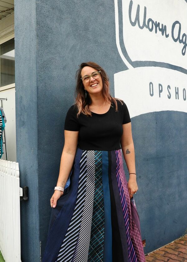 Woman standing in a skirt made of ties in front of a grey wall that says "worn again op shop"