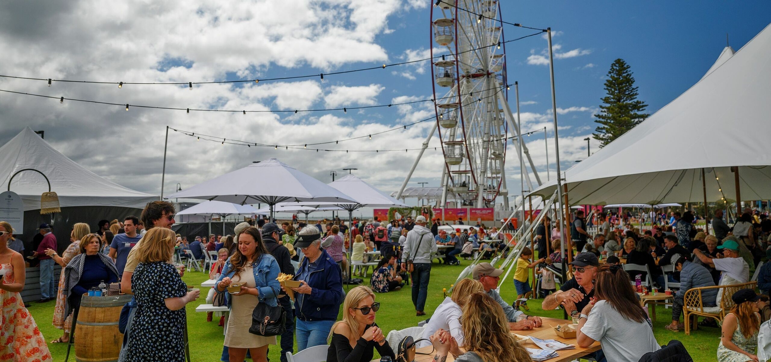 Large crowd of people standing, eating, and sitting near a ferris wheel