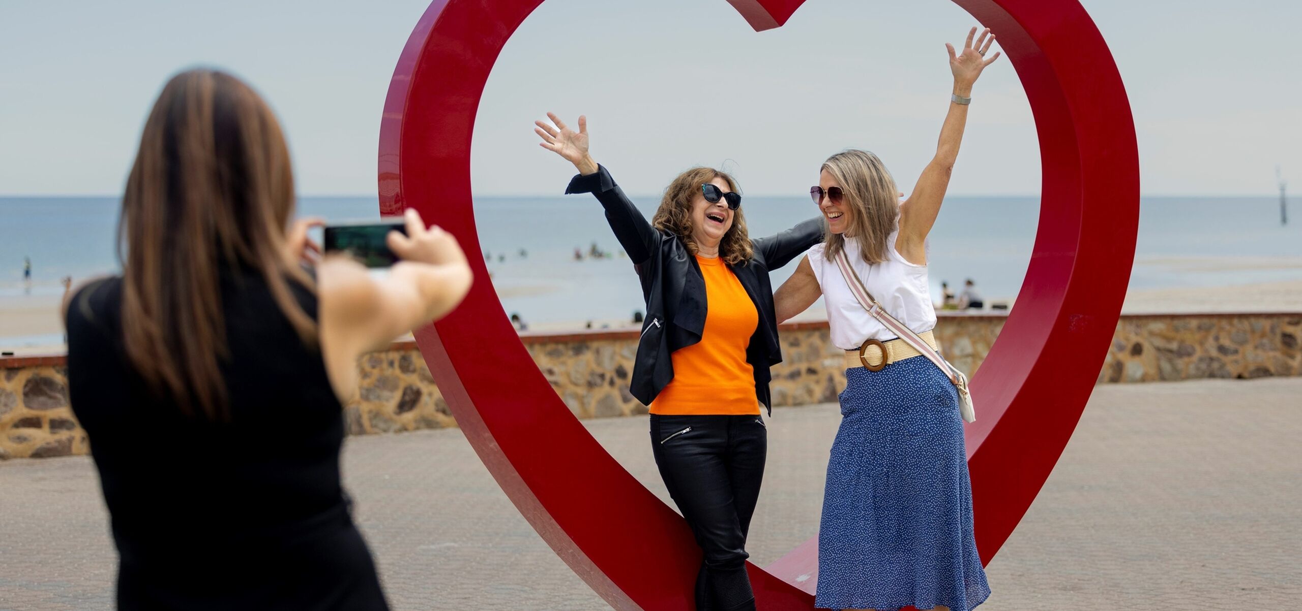 Two people posing in front of red heart sculpture. Other person taking a photo of it.