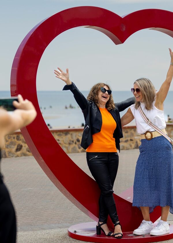 Two people posing in front of red heart sculpture. Other person taking a photo of it.
