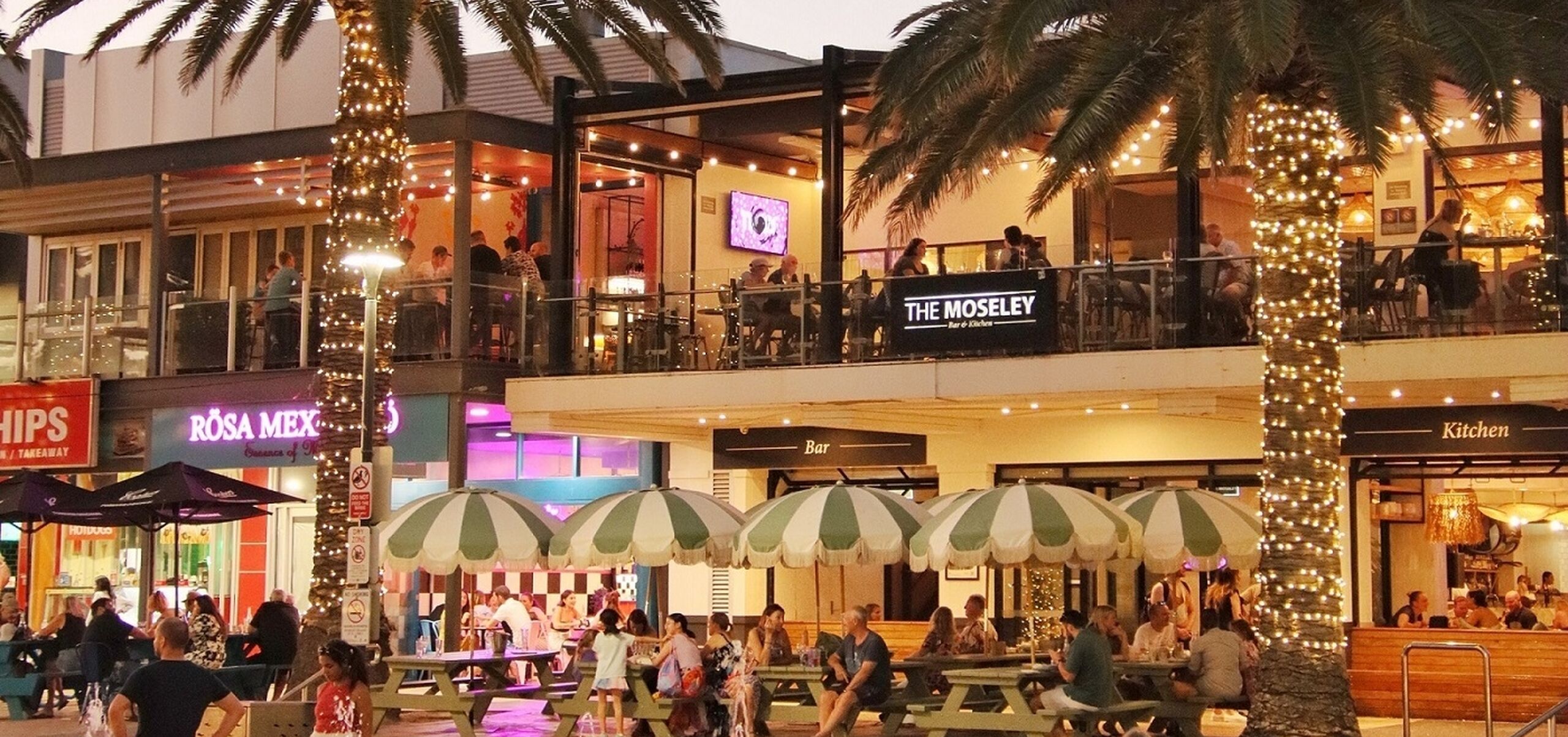 Children playing in water two palm trees and people sitting at outdoor dining tables