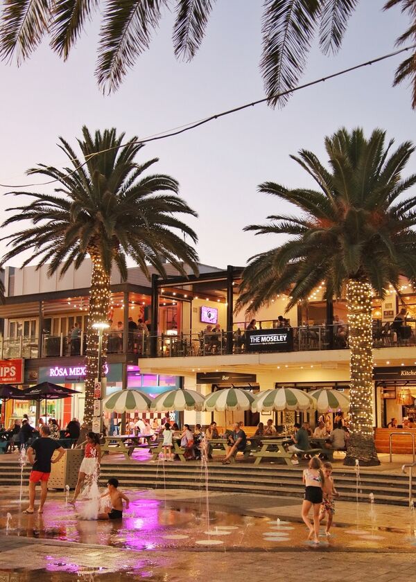 Children playing in water two palm trees and people sitting at outdoor dining tables