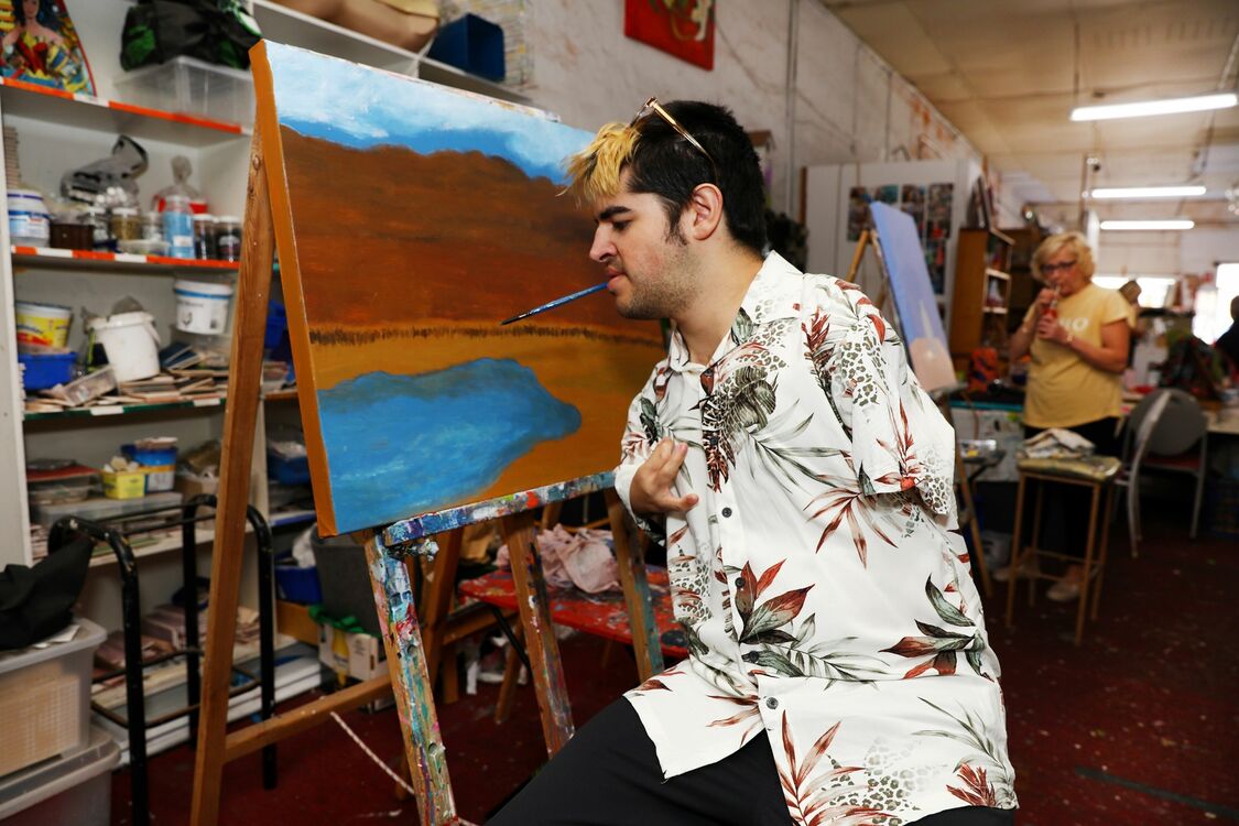 Person in white floral shirt painting with a paintbrush in their mouth