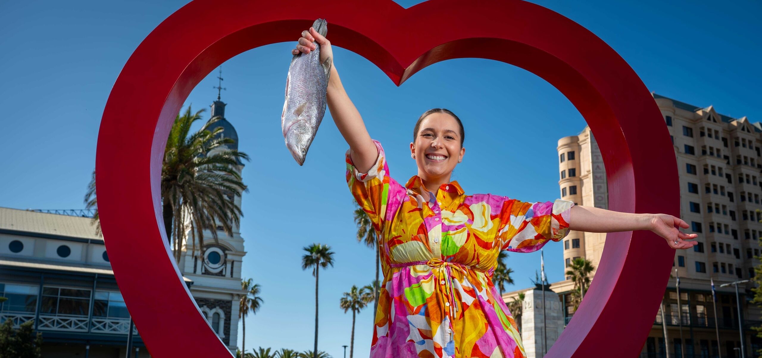 Laura Sharrad in bright dress holding fish in front of red heart in Glenelg