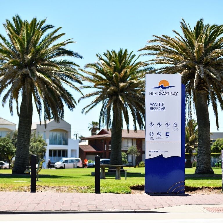 Reserve showing sign "City of Holdfast Bay. Wattle Reserve". Trees, paving and lawn area in scene.